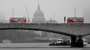 London bridge buses The photograph is an urban landscape taken in London, United Kingdom, during the early afternoon in winter. The image prominently features two iconic red London buses crossing a city bridge, set against the architectural backdrop of St Paul's Cathedral. The bridge, typical of London architecture, spans the River Thames and is surrounded by various riverboats and barges on the water. The misty, overcast atmosphere and bare trees in the scene suggest a cold winter's day. St Paul's Cathedral, a famous landmark, rises above the skyline and is visible through the haze, adding to the distinct London cityscape. The photograph captures the energy of city life and urban transportation in London, highlighting key elements such as the bridge, buses, and the historic cathedral in the heart of the United Kingdom's capital.